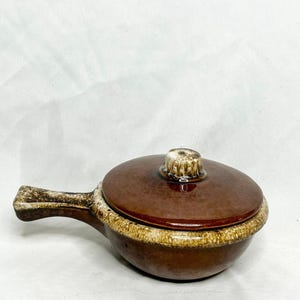 May include: A brown ceramic casserole dish with a lid and handle. The dish has a glossy finish and a two-tone design, with a darker brown lid and a lighter brown rim. The handle is a lighter brown color.