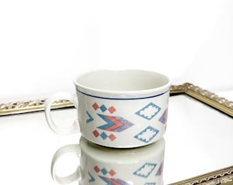May include: A white ceramic mug with a handle, featuring a geometric pattern in blue, red, and purple. The mug is set on a reflective surface, showcasing its mirrored image. The mug has a blue line near the rim.