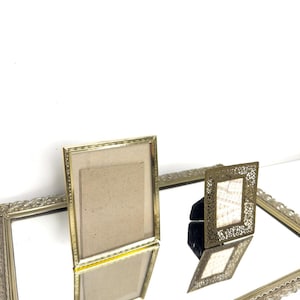 May include: Several antique gold-coloured picture frames, including a rectangular frame with a plain design and a smaller, ornate filigree frame. The frames are displayed on a mirrored surface.