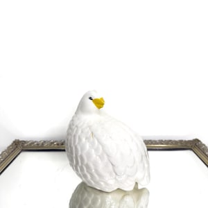 Può includere: Statuetta di anatra in ceramica bianca con becco giallo. L'anatra ha un aspetto strutturato e piumato ed è appoggiata su una superficie a specchio. La statuetta è un oggetto decorativo.