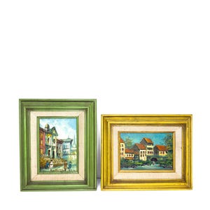 May include: Two framed landscape paintings. One painting is in a green frame and depicts a waterside scene with buildings and figures. The other painting is in a yellow frame and shows buildings with red roofs and a bridge.