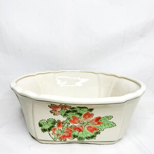 May include: Cream-colored ceramic planter with a scalloped rim and a floral design. The planter features a rectangular shape with a painted design of red flowers and green leaves. The rim is outlined with a black trim.