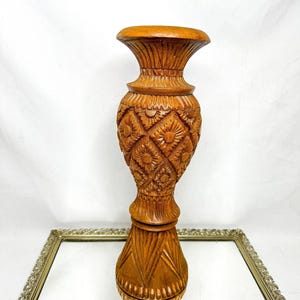 May include: A tall, intricately carved wooden vase with a warm, brown finish. The vase features detailed floral and geometric patterns, with a wide top and a flared base. The design includes carved diamond shapes and sunburst motifs, creating a decorative and artistic piece.