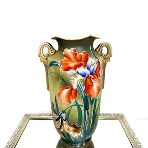 May include: An antique green vase with a floral iris design in red, white, and blue. The vase has gold-colored handles and a ruffled rim. The vase is displayed on a mirrored surface.