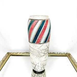 May include: A white ceramic vase with a cracked glaze effect. The upper portion features diagonal stripes in red, teal, and white. The vase is displayed on a reflective surface with a gold-colored frame.