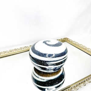 May include: A decorative, round ceramic object with a white and navy blue spiral design. The object is sitting on a mirrored surface within a gold-colored, ornate framed tray. The object's reflection is visible.