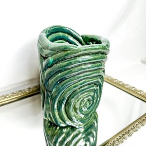 May include: A handcrafted, green ceramic vase with a unique spiral design. The vase has a glossy finish and is reflected in a mirrored surface. The vase is a decorative item.