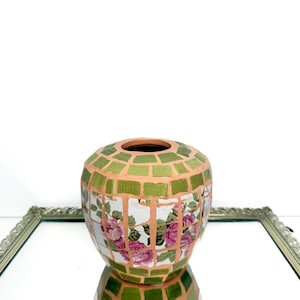 May include: A rounded vase featuring a mosaic design with green tiles and white tiles decorated with pink floral patterns. The tiles are separated by light orange grout lines, creating a grid-like appearance across the entire surface.