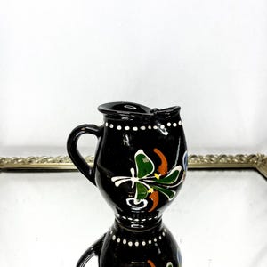 May include: A small, black ceramic pitcher with a handle and spout. The pitcher is decorated with colorful floral designs in green, orange, and blue. White dots encircle the top and bottom of the pitcher.