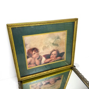 May include: A framed print featuring two cherubs with reddish-brown hair and dark wings against a cloudy sky. The artwork is set within a dark green mat and a gold-toned, textured frame with a rope-like border, reflecting in a mirror below.