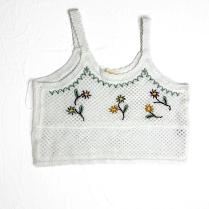 May include: White crochet crop top with a floral embroidery design. The top features a square neckline, spaghetti straps, and a scalloped edge. The embroidered flowers are yellow and brown.