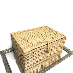 Puede incluir: Una cesta rectangular de mimbre tejido de color marrón claro con tapa con bisagras y cierre de cuerda. La cesta está sobre un espejo enmarcado plateado y ornamentado, que refleja su imagen. El fondo es blanco.