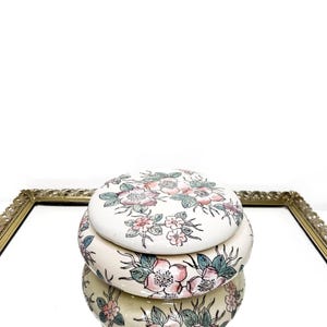May include: A stacked, round, ceramic trinket box with a lid. The box and lid are cream-colored and decorated with pink and purple floral designs and green leaves. The box is reflected in a mirror.