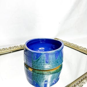 Può includere: Una ciotola in ceramica fatta a mano con un interno blu vibrante e un esterno strutturato in tonalità di blu e verde. La ciotola rotonda è posizionata su una superficie riflettente, mettendo in risalto la sua smaltatura e il suo design unici.