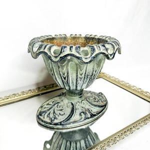 May include: A decorative, aged-looking, small, light green and black urn with a scalloped rim. The urn has an oval base with ornate detailing. The urn is sitting on a mirrored surface.
