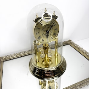 May include: A gold-toned anniversary clock with a circular face, black Roman numerals, and ornate filigree detailing. The clock is encased in a clear glass dome and features a rotating pendulum with four clear crystal-like spheres at the base.