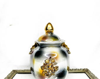 May include: A decorative ceramic jar with a lid, featuring a gold finial and floral embellishments. The jar has a color gradient of gray, white, yellow, and black. The jar is sitting on a mirrored surface.