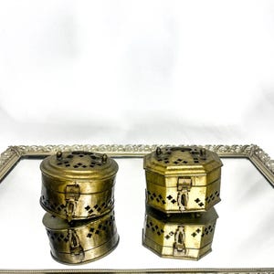 May include: Two antique brass boxes with intricate cut-out designs and hinged lids. The boxes are stacked on a mirrored surface within a decorative silver-toned frame. The boxes have a vintage aesthetic.