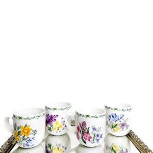 May include: Four white ceramic mugs with floral designs, including yellow daffodils, purple irises, and pink flowers. Each mug has a green vine border and is displayed on a gold-framed mirrored tray.