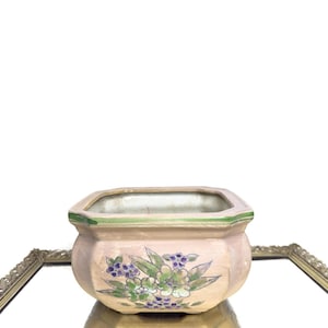 Peut inclure: Petit pot de fleurs octogonal en céramique rose clair, bordé de vert et orné de motifs floraux. Les fleurs sont violettes et blanches avec des feuilles vertes. Le pot se reflète dans un miroir.