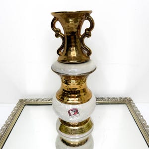 Puede incluir: Una botella decorativa vintage en forma de jarrón. Las secciones superior y media son doradas, mientras que el cuerpo es blanco con un patrón texturizado. La botella tiene un asa dorada y una etiqueta que dice "BEAM".