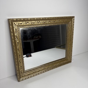 May include: A rectangular wall mirror featuring an ornate, textured gold-colored frame with intricate repeating patterns. The mirror surface reflects the surrounding environment, and a small black measuring tool is visible on the surface.