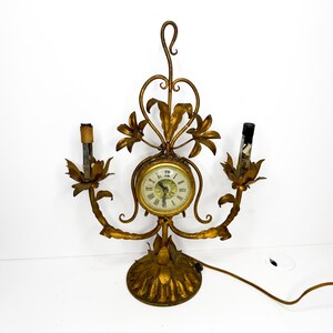 Può includere: Un orologio a candelabro in metallo dorato con un quadrante circolare centrale con numeri romani, affiancato da due portacandele a forma di fiore. La struttura presenta motivi ornamentali a foglie e viticci, una base circolare e un gancio.