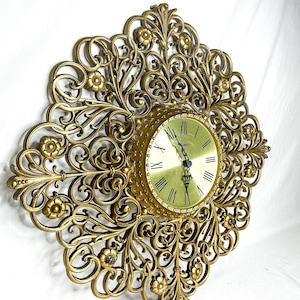 May include: A gold-colored wall clock with an ornate, floral filigree design. The clock face is round with Roman numerals and black hands. The clock's decorative frame features flowers and scrolling details.