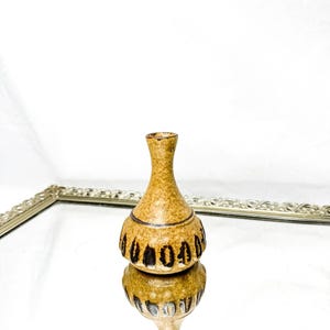 Puede incluir: Un pequeño jarrón de cerámica beige con base bulbosa y cuello estrecho. El jarrón presenta un patrón repetitivo marrón oscuro alrededor de la base. El jarrón se refleja en una superficie espejada, dentro de una bandeja plateada decorativa.