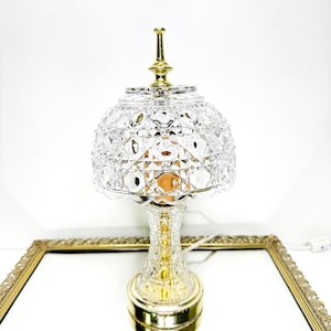 Könnte beinhalten: Eine klare Kristall-Tischlampe mit goldenem Sockel und Akzenten. Die Lampe hat einen facettierten Schirm und eine dekorative Spitze. Die Lampe steht auf einer verspiegelten Oberfläche in einem goldgerahmten Tablett.