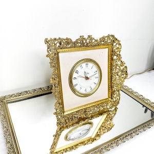 May include: A gold-toned, ornate square clock with a cream-colored face and a round clock face. The clock is set on a mirrored tray with a matching gold frame. The clock face has black numerals and red hands.