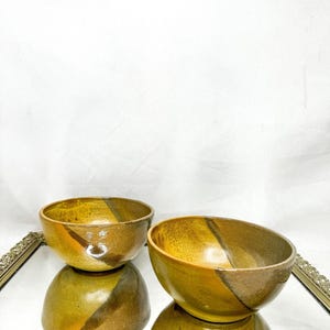 Può includere: Due ciotole in ceramica con finitura lucida. Le ciotole sono principalmente nei toni del marrone e del giallo, con una striscia diagonale di un colore marrone più scuro. Le ciotole sono su una superficie a specchio.