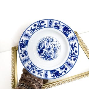 May include: White porcelain plate with a blue floral design. The plate features a central motif of birds and flowers, surrounded by a border of stylized floral patterns. The plate is held in front of a gold-framed mirror.