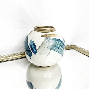 Può includere: Un piccolo vaso rotondo in ceramica con base bianca e disegni a pennellate blu e grigie. Il vaso ha un'apertura stretta e un bordo marrone. È esposto su una superficie riflettente.