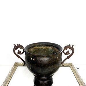 May include: A dark, weathered metal urn with a rounded body and a wide, open top. The vessel features two ornate, dragon-shaped handles on opposite sides and a fluted, pedestal-style base. The surface shows signs of oxidation and patina.