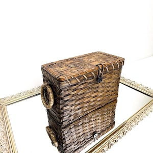 May include: A rectangular woven basket with a lid, featuring a button closure and two round handles. The basket is made of brown and tan woven material. The basket is sitting on a decorative mirror.