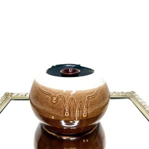 Puede incluir: Un cuenco decorativo de cerámica con exterior marrón y blanco, interior marrón oscuro y un pequeño acento redondo rojo oscuro. El cuenco se exhibe sobre una superficie espejada con un marco dorado decorativo.