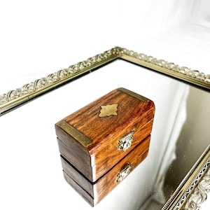 Pode incluir: Uma pequena caixa retangular de madeira com detalhes em latão e um emblema decorativo em latão na tampa. A caixa tem uma tampa articulada e um pequeno fecho. A caixa está sobre uma superfície espelhada.