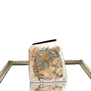 Puede incluir: Un jarrón de cerámica blanco con forma rectangular y parte superior inclinada. El jarrón presenta un diseño en relieve de hojas en tonos marrón y beige sobre un fondo azul claro. El jarrón se refleja en un espejo.