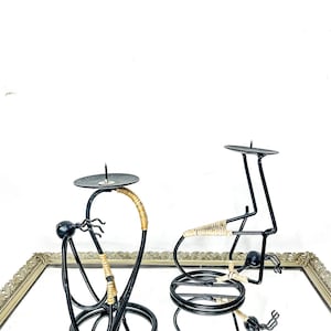 May include: Two black metal candle holders with a unique design. One is tall and curved, the other is shorter and more geometric. Both have a circular base and a small platform for a candle. The curved holder has a woven tan accent.