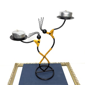 May include: A black metal candle holder with two figures, each holding a small, round candle. The figures have yellow and black accents, and the candles are silver. The candle holder is on top of a stack of blue books.