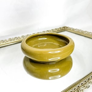 Könnte beinhalten: Eine kleine, runde, olivgrüne Keramikschale. Die Schale hat eine glatte, glänzende Oberfläche und steht auf einer reflektierenden Oberfläche, mit einem dekorativen goldfarbenen Rahmen im Hintergrund. Das schlichte Design der Schale macht sie für verschiedene Zwecke geeignet.