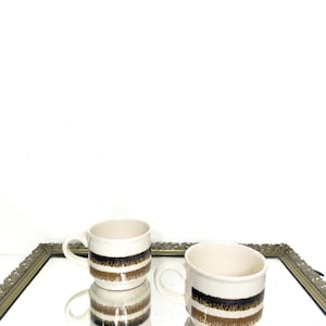 May include: Two cream-colored ceramic mugs with a brown and black stripe design. The mugs have a handle and are displayed on a mirrored surface. The mugs are part of a coffee or tea set.