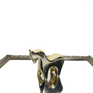 May include: A gold-colored horse figurine with a cream-colored saddle. The horse is reflected in a mirrored surface, set within a silver-toned, ornate frame. The background is white.