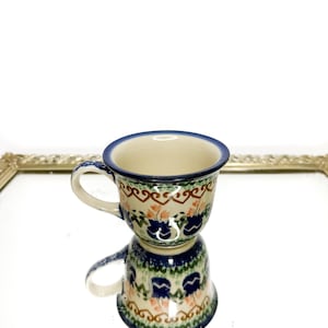 Może przedstawiać: Ceramiczny kubek o kremowej podstawie z ręcznie malowanym wzorem kwiatowym w odcieniach niebieskiego, zielonego i pomarańczowego. Kubek posiada ciemnoniebieski brzeg, zakrzywione ucho oraz ozdobną brązową linię w zygzak w górnej części.