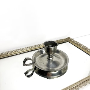 Puede incluir: Un candelero vintage de color plateado con asa. El candelero tiene una base redonda y una copa para la vela. El asa tiene un diseño de bucle. El candelero está sobre una superficie espejada.