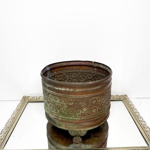 May include: A round, weathered copper planter featuring an embossed floral pattern around the exterior. The vessel has a dark, oxidised patina with green verdigris accents and rests on small, integrated feet, showcasing a textured metallic surface.