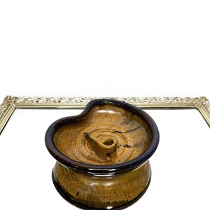 May include: A handmade ceramic candle holder with a brown and blue glaze. The candle holder has a unique, organic shape with a central well for the candle and a curved rim. The piece is displayed on a mirrored surface.