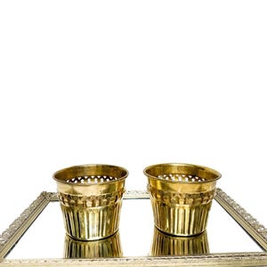 Könnte beinhalten: Zwei kleine, goldfarbene Metallpflanzgefäße mit einem dekorativen Lochmuster. Die Pflanzgefäße sind zylindrisch und stehen auf einer verspiegelten Oberfläche in einem rechteckigen, verzierten Tablett. Der Hintergrund ist weiß.