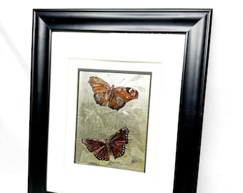 May include: Framed artwork featuring two detailed butterfly illustrations. The butterflies have brown, orange, and black wings, set against a muted green and gray background. The artwork is framed in a black wooden frame with a white mat.